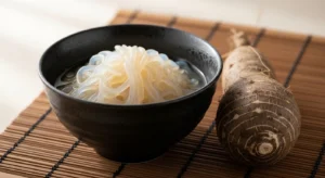 Fideos shirataki translucidos de konjac en bowl de ceramica oscura junto a raiz de konjac sobre esterilla de bambu